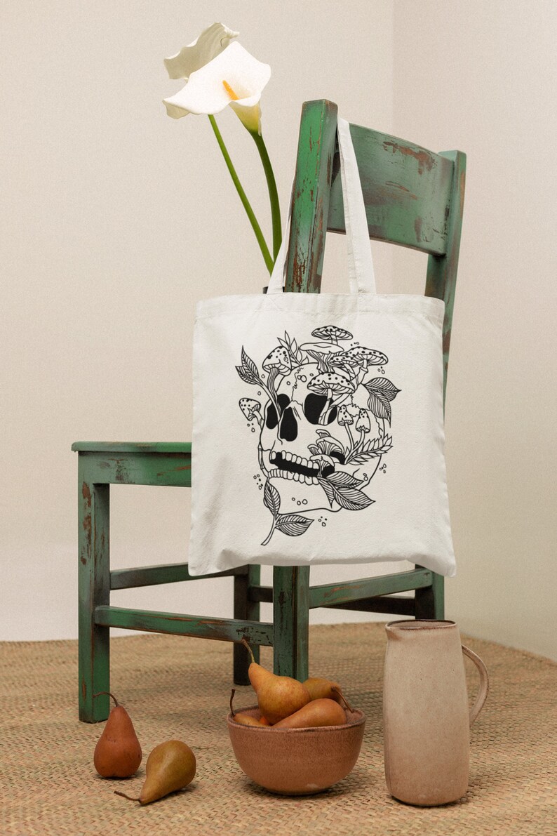 May include: White tote bag with a black line drawing of a skull with mushrooms growing out of it. The bag is sitting on a green chair with a white flower in the background.