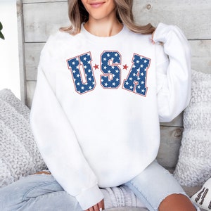 Puede incluir: Sudadera blanca con un gr&aacute;fico "USA" azul y rojo con estrellas blancas. Las letras est&aacute;n delineadas en rojo.