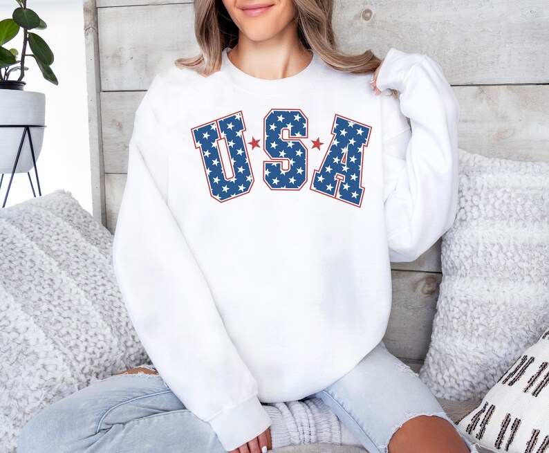 Puede incluir: Sudadera blanca con un gr&aacute;fico "USA" azul y rojo con estrellas blancas. Las letras est&aacute;n delineadas en rojo.