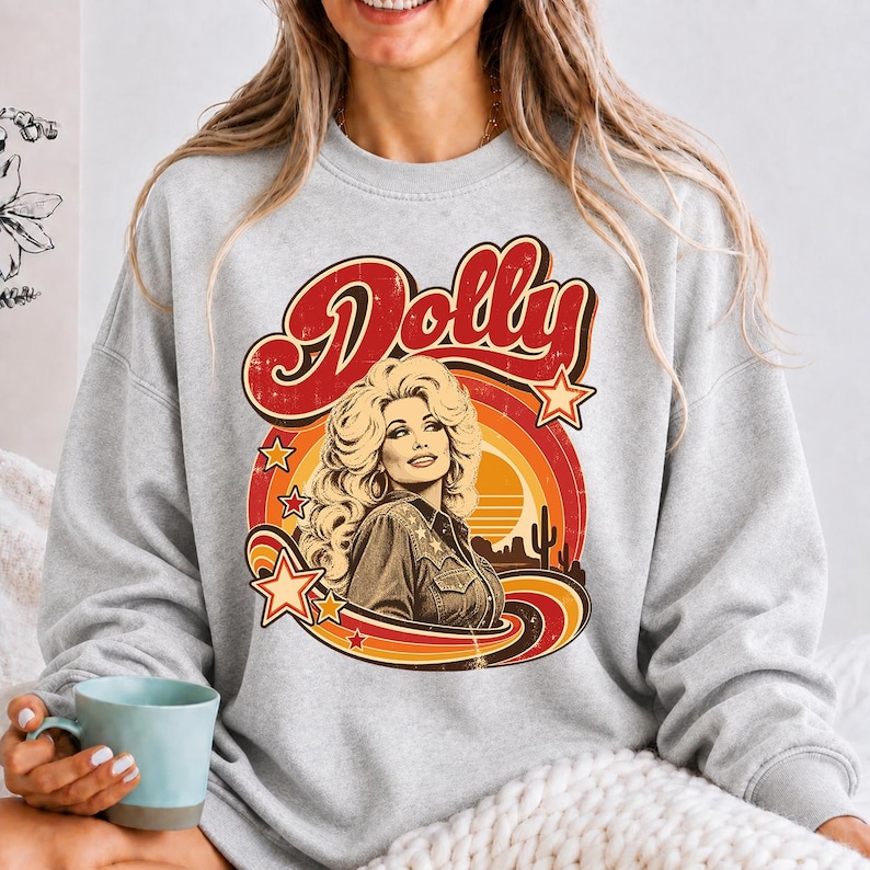 May include: A light gray crewneck sweatshirt featuring a graphic print of a blonde woman with voluminous hair wearing a collared shirt. The design includes the word Dolly in red script, orange and yellow circular sun rays, and small stars.