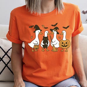 Pu&ograve; includere: Maglietta arancione con un motivo a tema Halloween. Il design include quattro anatre bianche vestite con costumi di Halloween, tra cui cappelli da strega e una zucca. Un gatto nero e dei pipistrelli fanno parte del design.