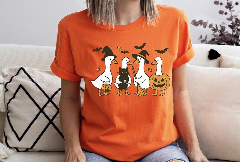Pu&ograve; includere: Maglietta arancione con un motivo a tema Halloween. Il design include quattro anatre bianche vestite con costumi di Halloween, tra cui cappelli da strega e una zucca. Un gatto nero e dei pipistrelli fanno parte del design.