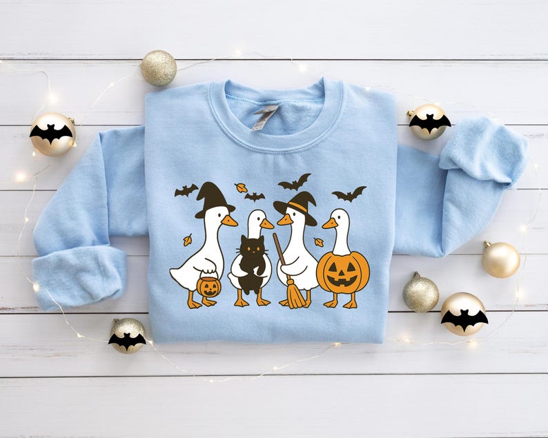 Pu&ograve; includere: Una felpa azzurra con un motivo a tema Halloween. Il design include quattro anatre bianche vestite con cappelli da strega e che tengono in mano una zucca, una scopa e un secchio per dolcetti. &Egrave; presente anche un gatto nero.