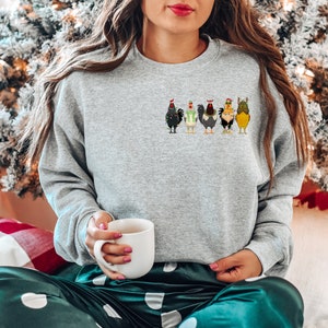 May include: A grey sweatshirt featuring a festive design of five chickens wearing Christmas hats and jumpers. The sweatshirt is being worn by a person sitting on a green and white patterned blanket.