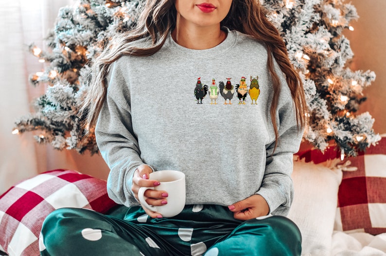 May include: A grey sweatshirt featuring a festive design of five chickens wearing Christmas hats and jumpers. The sweatshirt is being worn by a person sitting on a green and white patterned blanket.