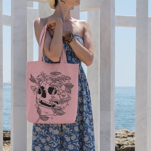 May include: A pink tote bag with a black and white illustration of a skull with mushrooms growing out of it. The bag is being held by a person wearing a floral dress.