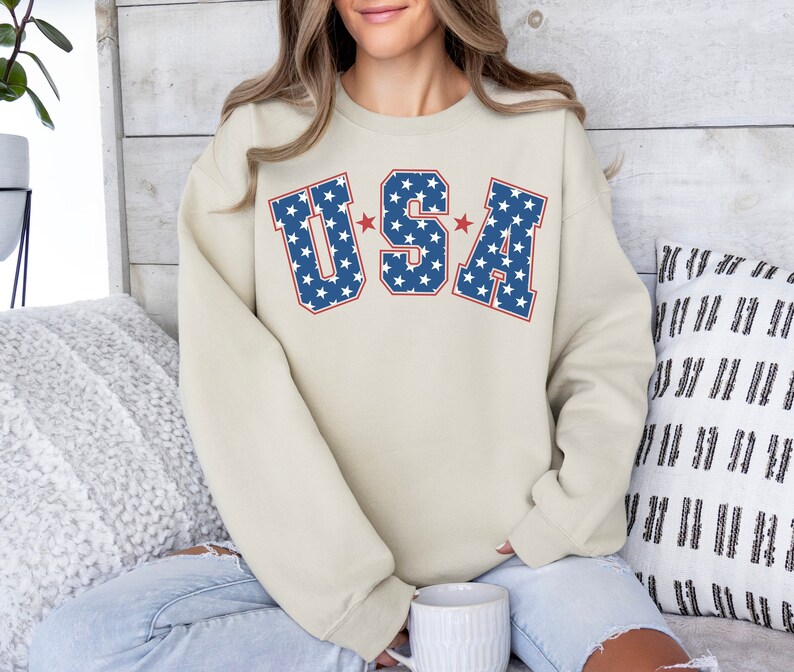 Puede incluir: Una sudadera beige con un gr&aacute;fico "USA" azul y rojo con estrellas blancas. El gr&aacute;fico est&aacute; en una fuente de estilo universitario.