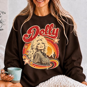 May include: A dark brown long-sleeve sweatshirt featuring a graphic print of a woman with voluminous blonde hair. The design includes the word Dolly in red script, a sunset motif with orange and yellow rings, stars, and a desert landscape.