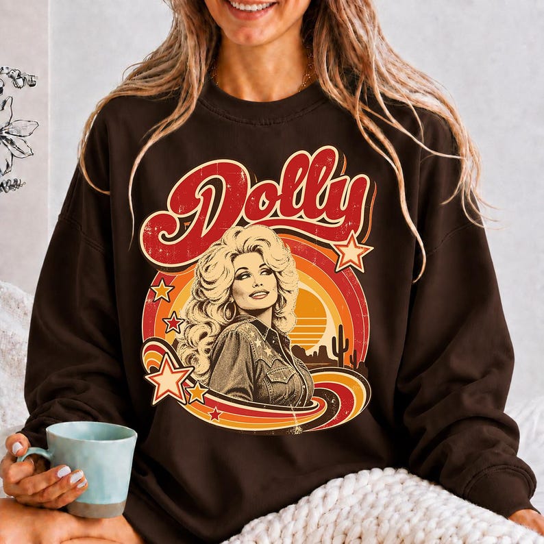 May include: A dark brown long-sleeve sweatshirt featuring a graphic print of a woman with voluminous blonde hair. The design includes the word Dolly in red script, a sunset motif with orange and yellow rings, stars, and a desert landscape.