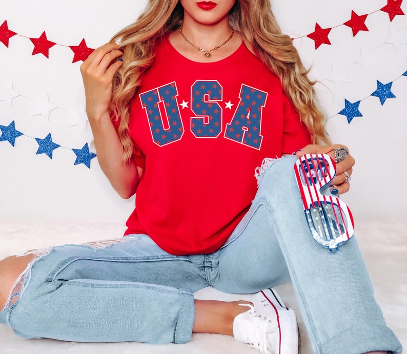 Puede incluir: Una camiseta roja con la palabra "USA" impresa en azul y estrellas blancas. La camiseta la lleva puesta una persona con el pelo largo y rubio y unos vaqueros azules. La persona sostiene unas gafas rojas, blancas y azules.