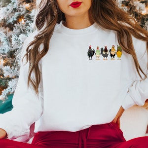 May include: White sweatshirt featuring a festive design of six chickens wearing Santa hats. The chickens are arranged in a line, facing forward, and each has a unique colour and pattern.