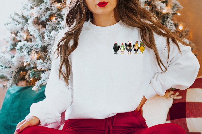 May include: White sweatshirt featuring a festive design of six chickens wearing Santa hats. The chickens are arranged in a line, facing forward, and each has a unique colour and pattern.