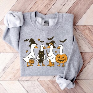 Pu&ograve; includere: Una felpa grigia con un design a tema Halloween. La grafica mostra quattro oche dei cartoni animati con cappelli da strega che tengono in mano una zucca, un gatto nero e una scopa. Il design include anche pipistrelli e foglie.