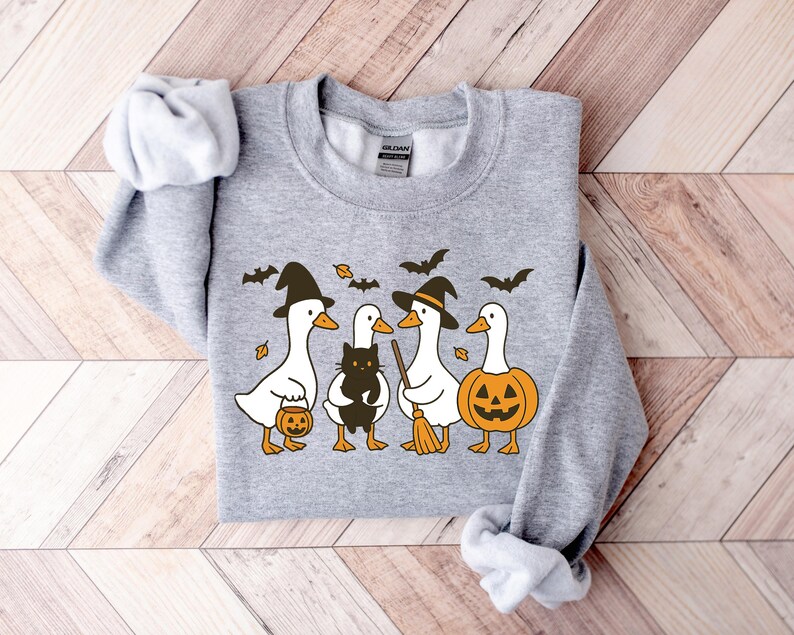Pu&ograve; includere: Una felpa grigia con un design a tema Halloween. La grafica mostra quattro oche dei cartoni animati con cappelli da strega che tengono in mano una zucca, un gatto nero e una scopa. Il design include anche pipistrelli e foglie.