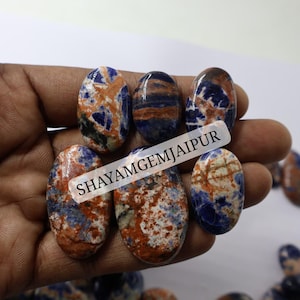 May include: A collection of oval-shaped cabochons featuring a mottled pattern of deep blue, rusty orange, and white mineral inclusions. A semi-transparent label with the black text SHAYAMGEMJAIPUR is overlaid across the center of the stones.