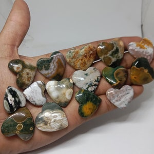 May include: A collection of 14 heart-shaped polished stones with various patterns of green, brown, white, and yellow. The stones are arranged on a hand.