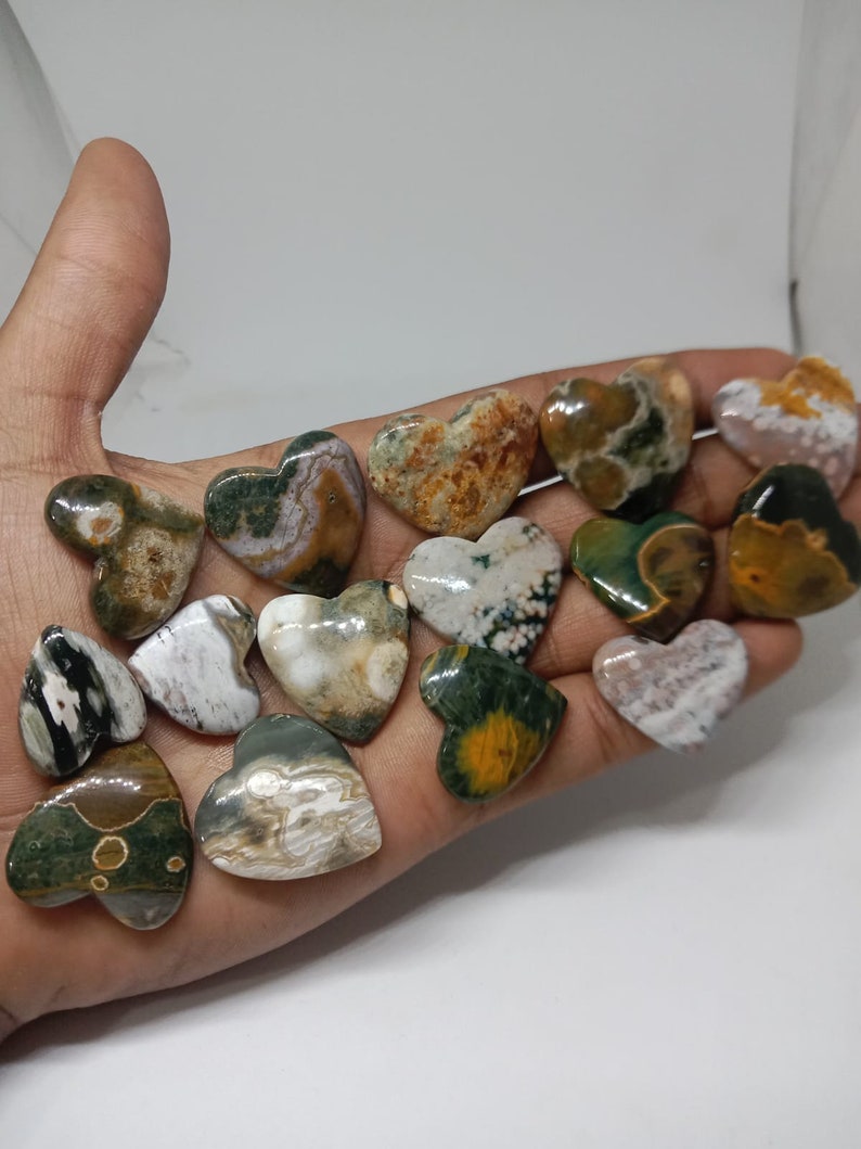May include: A collection of 14 heart-shaped polished stones with various patterns of green, brown, white, and yellow. The stones are arranged on a hand.