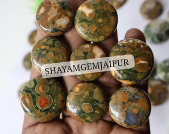 Puede incluir: Una colección de cabujones de jaspe orbicular redondos y pulidos con un patrón moteado de inclusiones circulares en verde bosque, bronceado y naranja quemado. Una etiqueta semitransparente con el texto negro SHAYAMGEMJAIPUR está centrada sobre las piedras.