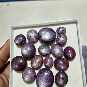 Puede incluir: Una colecci&oacute;n de 15 cabujones de rub&iacute; estrella p&uacute;rpura en una caja blanca. Las piedras son de forma ovalada y tienen un patr&oacute;n de estrella.