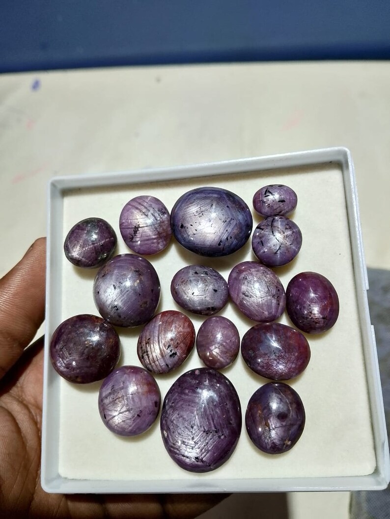 Puede incluir: Una colecci&oacute;n de 15 cabujones de rub&iacute; estrella p&uacute;rpura en una caja blanca. Las piedras son de forma ovalada y tienen un patr&oacute;n de estrella.