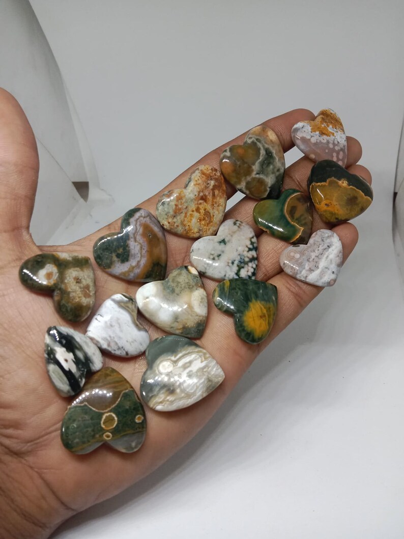 May include: A collection of 15 heart-shaped polished stones with green, brown, and white patterns. The stones are arranged in a hand.