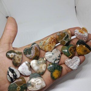 May include: A collection of 15 heart-shaped polished stones with a variety of colors and patterns, including green, brown, white, and black. The stones are arranged in a hand.