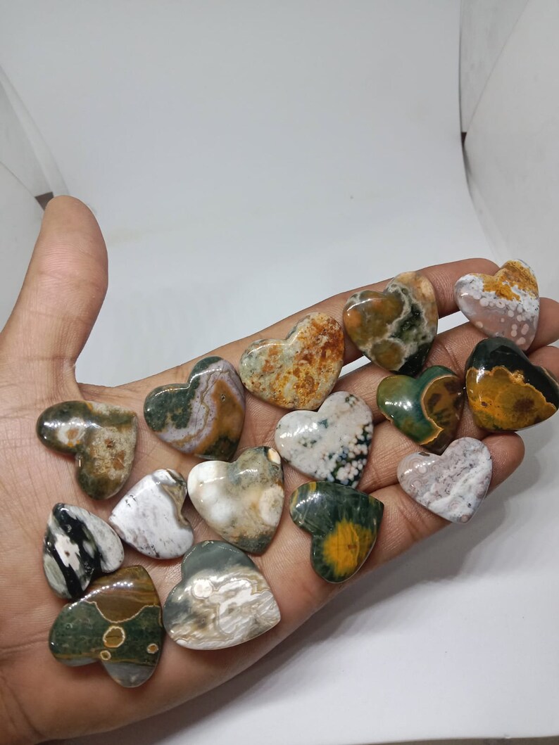 May include: A collection of 15 heart-shaped polished stones with a variety of colors and patterns, including green, brown, white, and black. The stones are arranged in a hand.