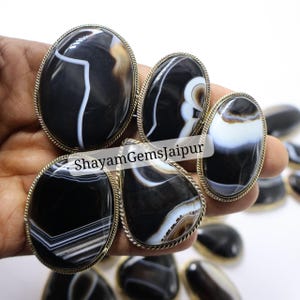 May include: A selection of black and white agate gemstones in oval and teardrop forms. Each stone is set in a silver-coloured metal bezel with a rope-like edge. The gemstones are displayed against a light background.