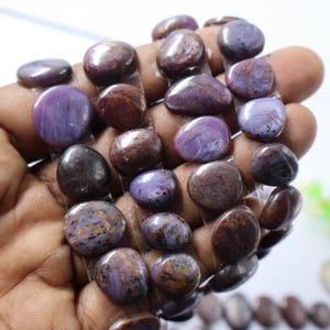 Puede incluir: Una colección de piedras preciosas pulidas, de forma ovalada, en tonos morados y marrones. Las piedras están ensartadas, mostrando sus superficies lisas y redondeadas y sus variaciones de color naturales. Las piedras se sostienen en una mano.