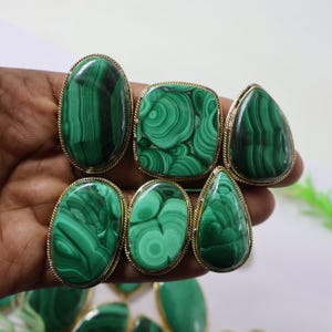 May include: A selection of malachite rings with gold-coloured settings. The rings display diverse shapes, such as oval, square, and teardrop, highlighting the stone's unique green and black patterns. The rings are presented against a neutral backdrop.