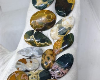 May include: A collection of 11 polished ocean jasper stones in various shapes and sizes. The stones feature a mix of green, brown, yellow, and white colors with intricate patterns.