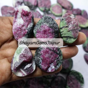Può includere: Una collezione di gemme di rubino grezze con una miscela di colori, tra cui rosso intenso, nero, verde e bianco. Le gemme hanno una consistenza naturale e ruvida e forme diverse. Il testo "ShayamGemsJaipur" è visibile.