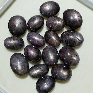 May include: A collection of oval-shaped, dark purple star sapphires. Each stone displays a prominent star effect, with lines radiating from a central point. The gems have a polished, reflective surface, and are arranged on a light-colored surface.