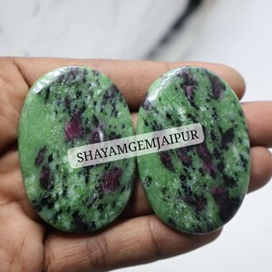 May include: Two oval-shaped polished ruby zoisite gemstones featuring a vibrant green base with speckled black inclusions and patches of deep magenta-red. A white rectangular label with the black text SHAYAMGEMJAIPUR is centered across both stones.