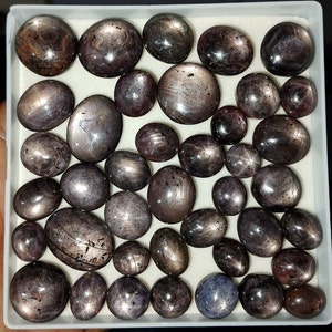 May include: A collection of 30+ oval-shaped, dark gray star sapphires with a silver sheen. The stones are arranged in a white box.