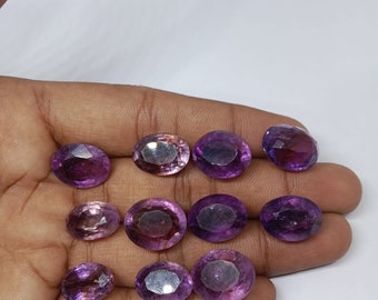 Könnte beinhalten: Eine Sammlung von zehn ovalen Amethyst-Edelsteinen in verschiedenen Violetttönen. Die Edelsteine sind in einem Halbkreis auf einer Hand angeordnet.
