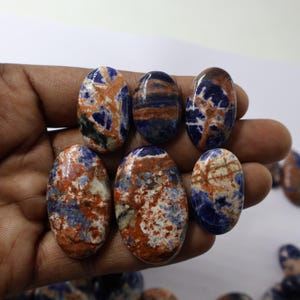 May include: Six oval-shaped sodalite gemstones with a mix of blue, orange, and white patterns. The stones are held in a hand, showcasing their polished surfaces and natural color variations. The gemstones are ideal for jewelry making.