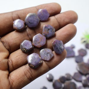 May include: A collection of rough, hexagonal-shaped gemstones in shades of purple and brown. The gemstones are held in a hand, showcasing their natural, unpolished texture and unique color variations. These stones are likely used for jewelry or collecting.