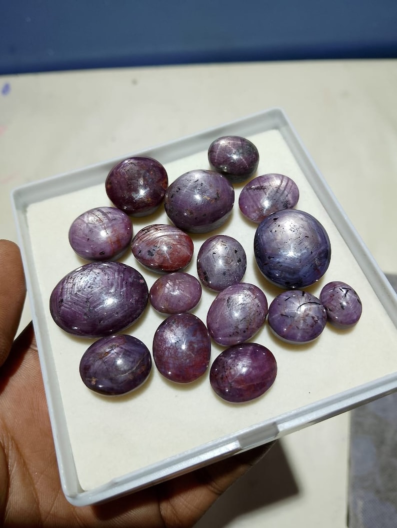 Puede incluir: Una colecci&oacute;n de 15 piedras preciosas de rub&iacute; estrella p&uacute;rpura en una caja blanca. Las piedras preciosas son de forma ovalada y redonda y tienen un patr&oacute;n de estrella.