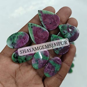 May include: A collection of polished ruby zoisite gemstones in various oval and teardrop shapes, featuring a mix of vibrant magenta and deep green colors with black speckles. The text SHAYAMGEMSJAIPUR is printed across the center of the stones.