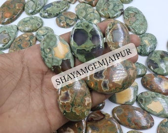 Puede incluir: Una colección de cabujones pulidos de forma ovalada con un patrón moteado en colores verde, marrón y bronceado. Las piedras tienen un acabado liso y brillante. Una etiqueta rectangular blanca con el texto negro SHAYAMGEMJAIPUR está colocada.