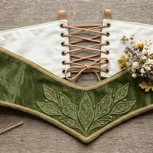 May include: A wide olive green velvet corset belt featuring a cream-colored interior, gold braided trim, and a central front panel embroidered with leaf motifs. The back includes a tan leather lace-up closure with metal grommets.