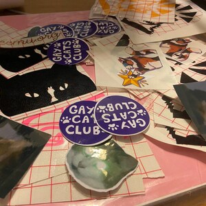 May include: A collection of stickers featuring black cats, a rainbow cat, and the text "Gay Cats Club". The stickers are on a pink and white gridded surface.