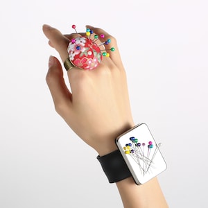 Può includere: Un cuscino ad anello floreale rosso con spilli colorati e un cuscino da polso abbinato con cinturino in pelle nera. Il cuscino ad anello è decorato con un motivo floreale e il cuscino da polso ha uno sfondo bianco.