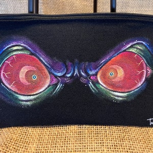 May include: Black canvas zippered pouch with a hand-painted design of two large, red eyes with purple and green accents. The pouch is signed "TMJ 23".
