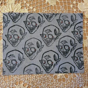 May include: A rectangular gray fabric with a repeating pattern of grinning, cartoonish faces with red eyes. The faces have pointed ears and are outlined in black. The fabric is likely a decorative item.