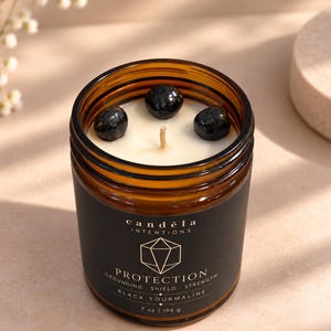 May include: A candle in an amber glass jar with a black label reading candela intentions PROTECTION GROUNDING SHIELD STRENGTH BLACK TOURMALINE 7 oz | 198 g. The white wax contains three smooth, round black tourmaline stones and a single central wick.