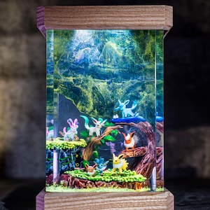 May include: A rectangular, illuminated resin diorama featuring a miniature forest scene with various colourful cartoon creatures. The diorama is encased in a wooden frame and sits on a dark surface. A small house-shaped object is visible in the background.