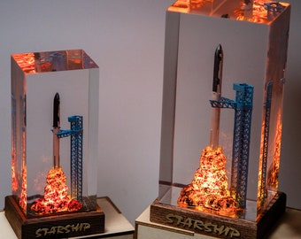 May include: Two illuminated resin sculptures depicting a rocket launch. The rockets are black, with blue launch structures, and fiery orange bases. The sculptures are encased in clear rectangular blocks, mounted on wooden bases with the word "STARSHIP".
