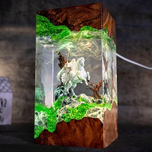 May include: A white resin figurine of a person riding a horse is encased in a clear resin block. The block is set in a wooden base with green moss and a tree branch. The resin block is illuminated from within, creating a glowing effect.